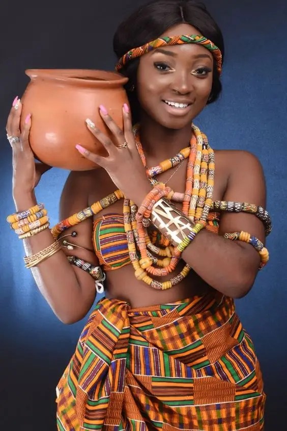 A woman wearing beaded kente fabric holding a clay pot