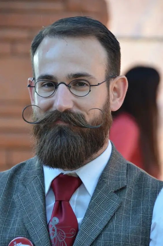 A man wearing glasses has a beard and a unique mustache