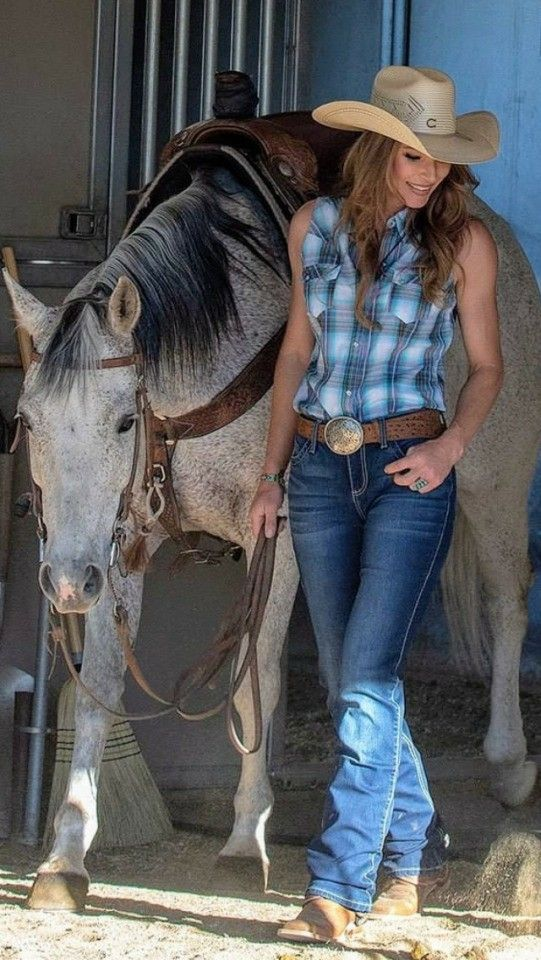 A cowgirl wearing a sleeveless plaid shirt, hat and jeans standing by her horse