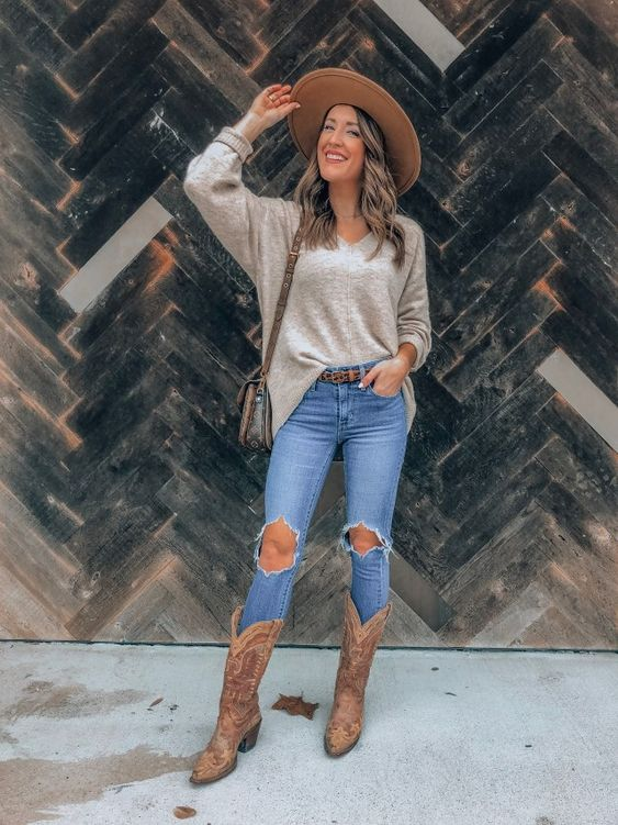 Smiling woman shows cowgirl outfit ideas with jeans, sweater, hat and sling bag